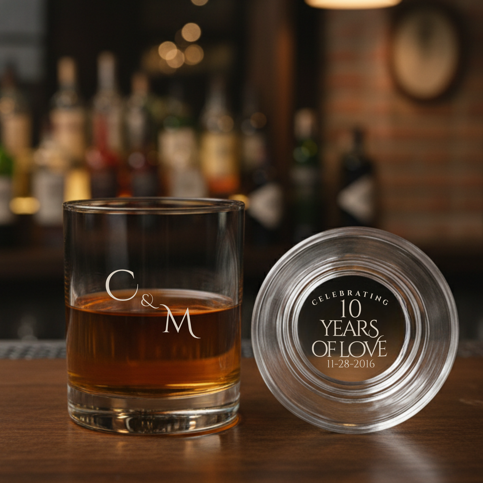 Personalized whiskey glass engraved with sample initials "C&M" on front and bottom engraving reading "Celebrating 10 Years of Love 11-28-2016," filled with amber liquid, shown with matching coaster