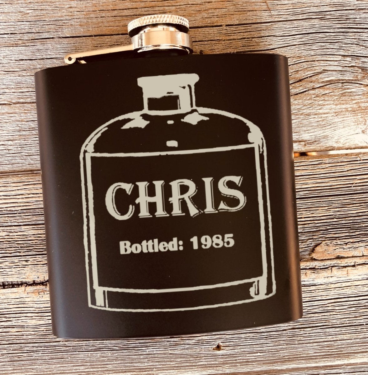 Black flask on weathered wood engraved with a vintage bottle label reading CHRIS and Bottled: 1985, a unique birthday gift for him with his year