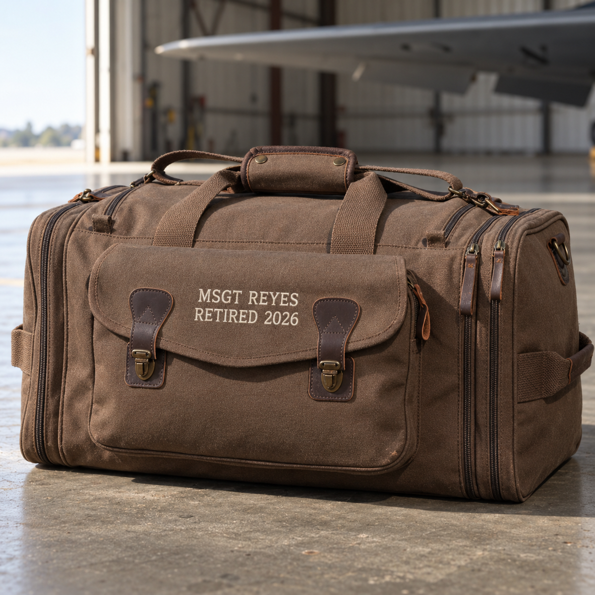 Brown canvas Air Force duffle bag with leather trim embroidered MSGT REYES and RETIRED 2026 in ivory thread, on an aircraft hangar floor, a personalized retirement gift