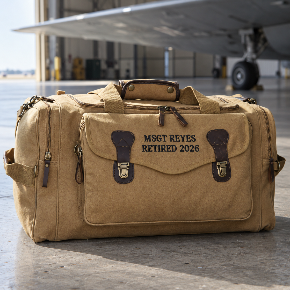 Tan canvas Air Force duffle bag with leather trim embroidered MSGT REYES and RETIRED 2026, standing on an aircraft hangar floor, a personalized retirement gift