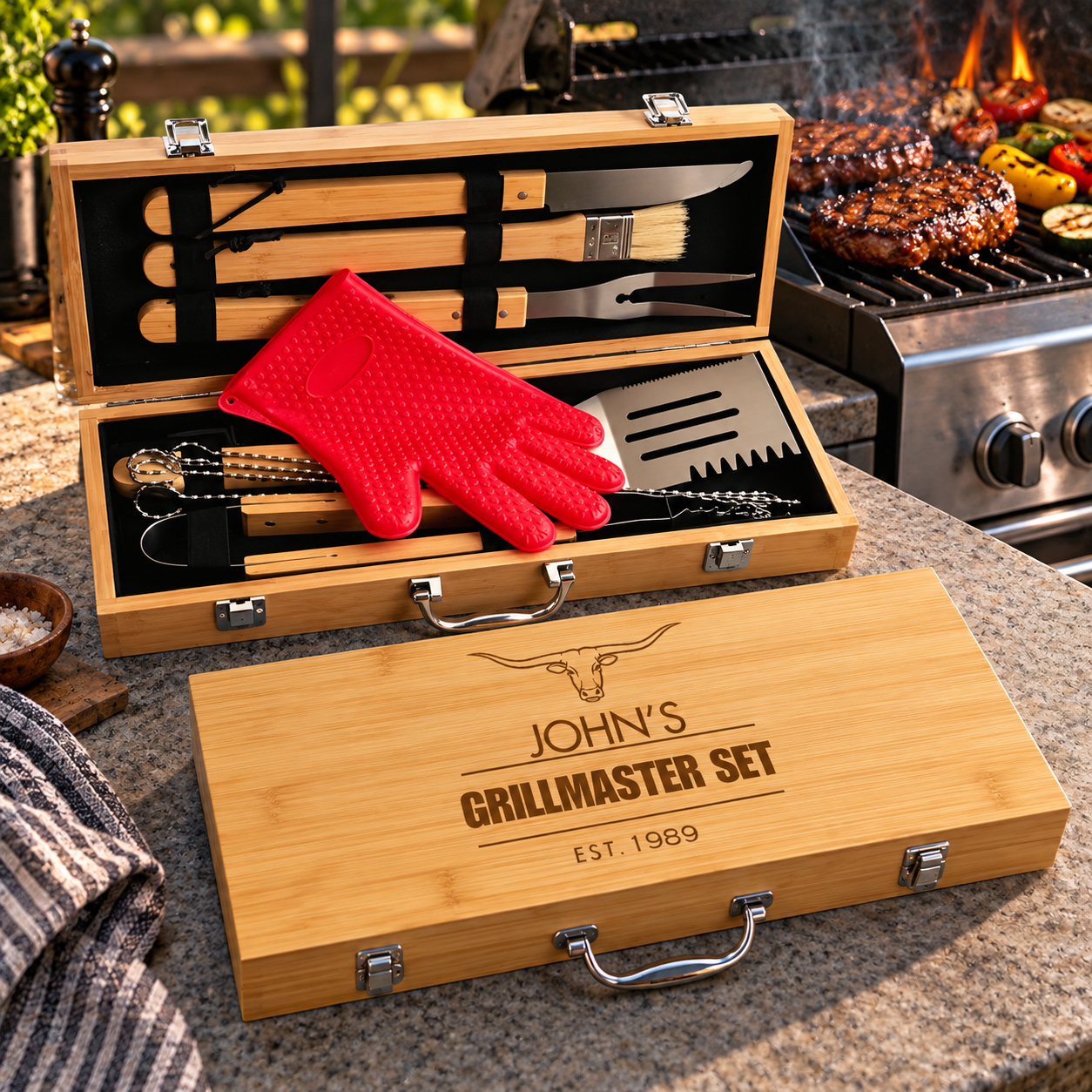 Two wooden grill tool boxes with engraved "JOHN'S GRILLMASTER SET EST 1989" on lower box, containing spatulas, tongs, forks, and red grilling glove beside a lit grill.