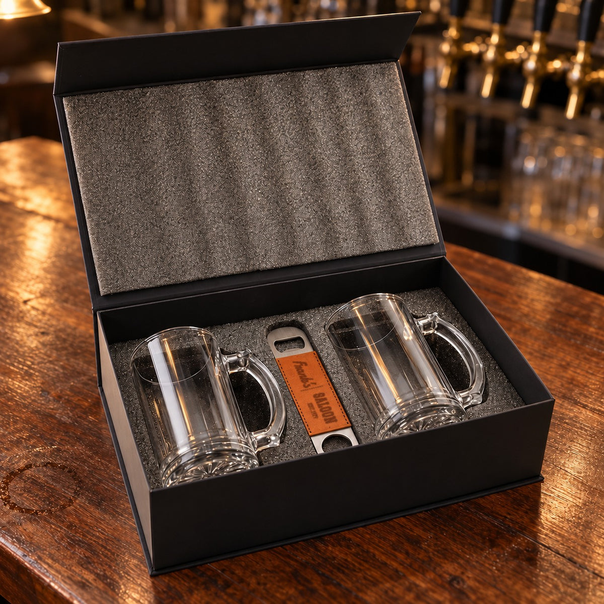 Beer mug gift set open in its foam-lined magnetic box: two clear glass beer mugs and a leather bottle opener engraved Frank's Saloon Est. 1979