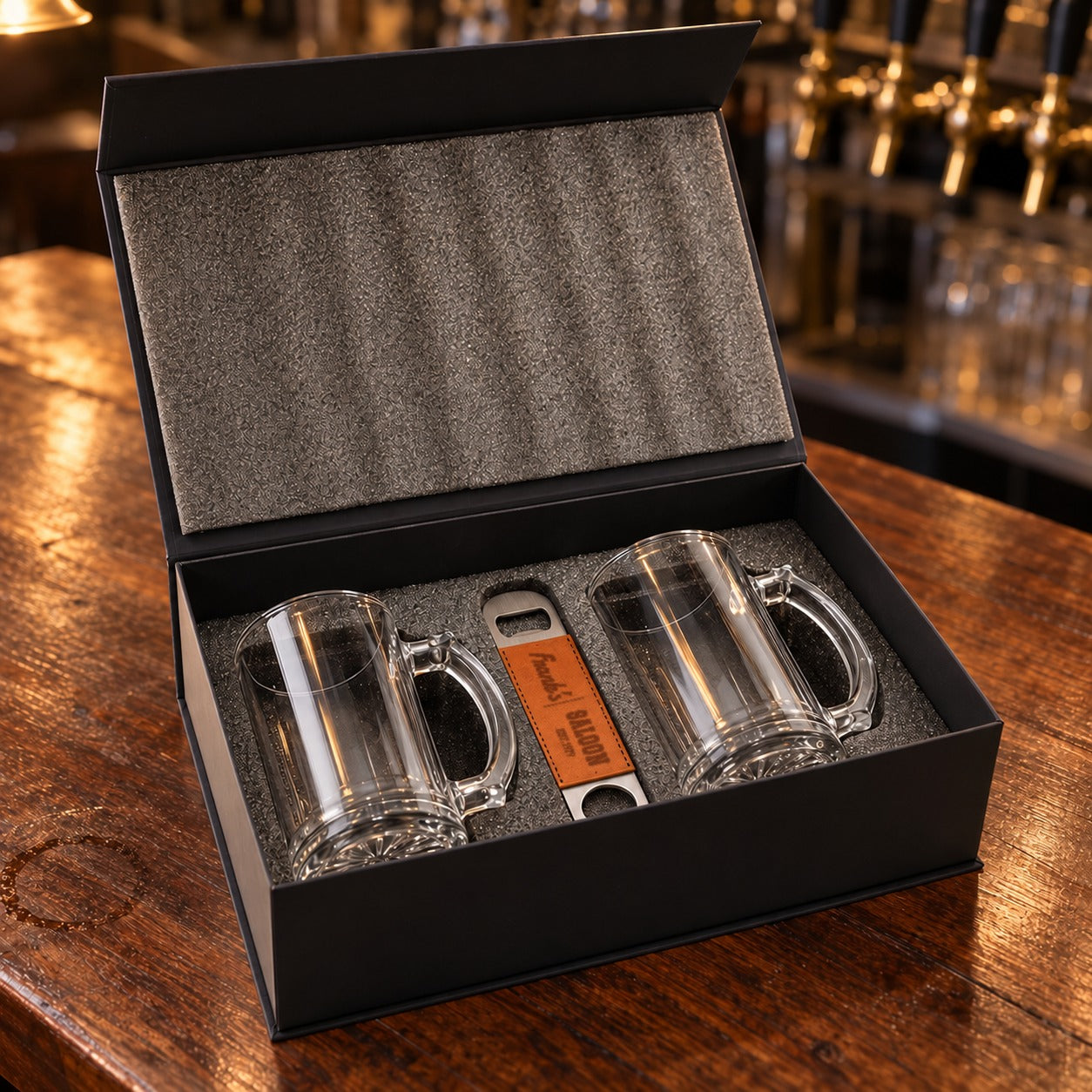 Beer mug gift set open in its foam-lined magnetic box: two clear glass beer mugs and a leather bottle opener engraved Frank's Saloon Est. 1979