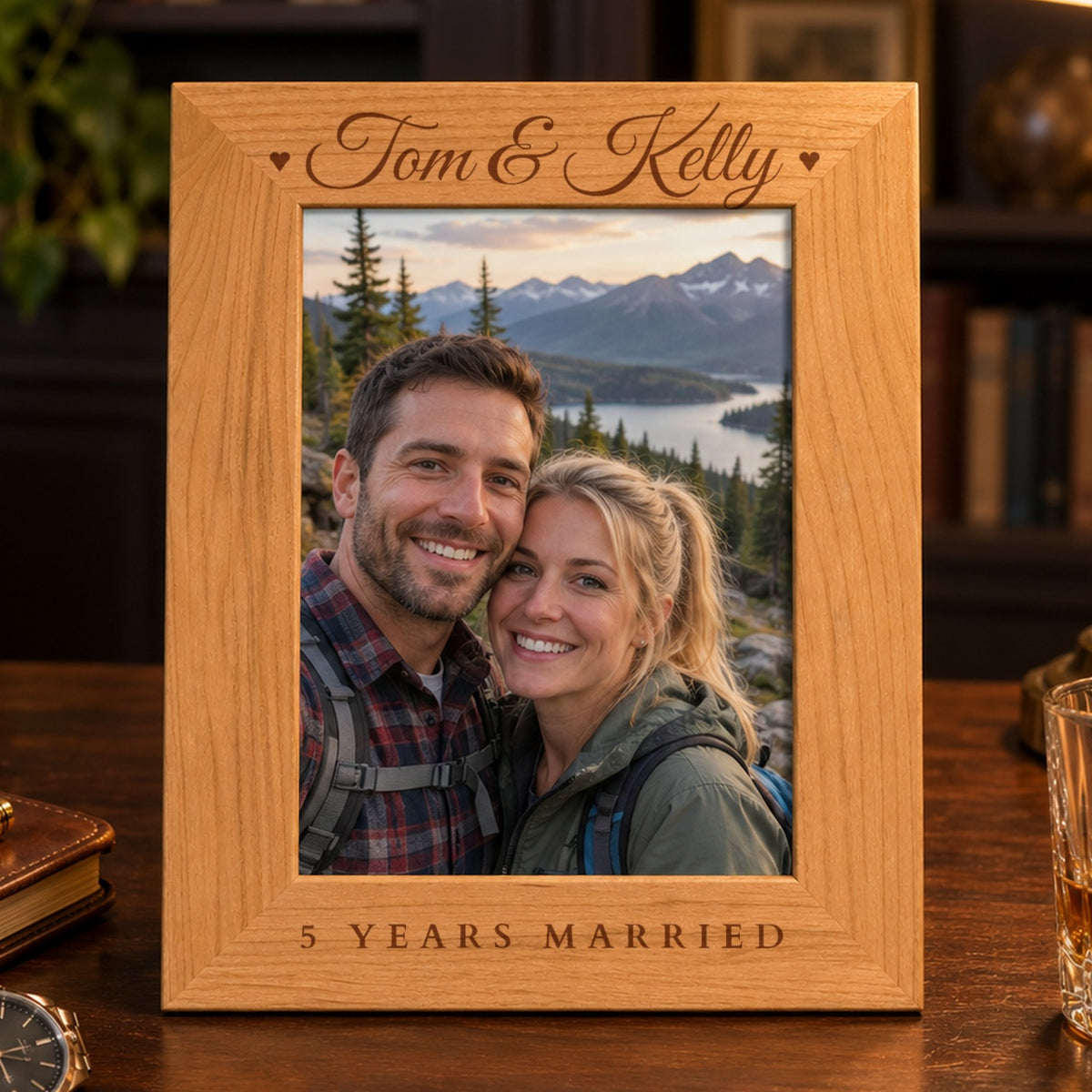 Personalized picture frame in red alder engraved Tom & Kelly with hearts and 5 Years Married, holding a hiking photo on a walnut desk beside a watch