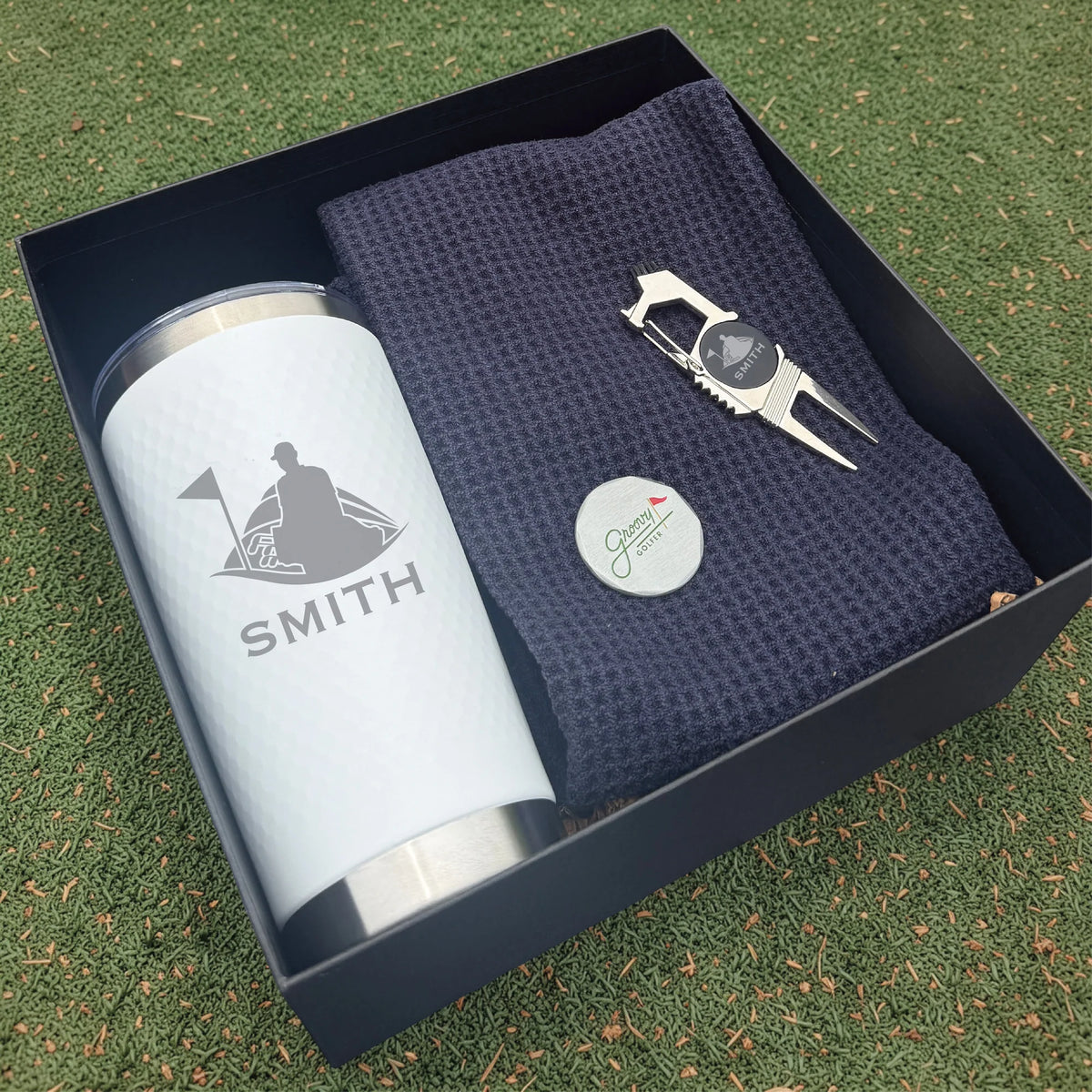 Golf-themed gift set with tumbler, towel, and divot tool in a box on grass.