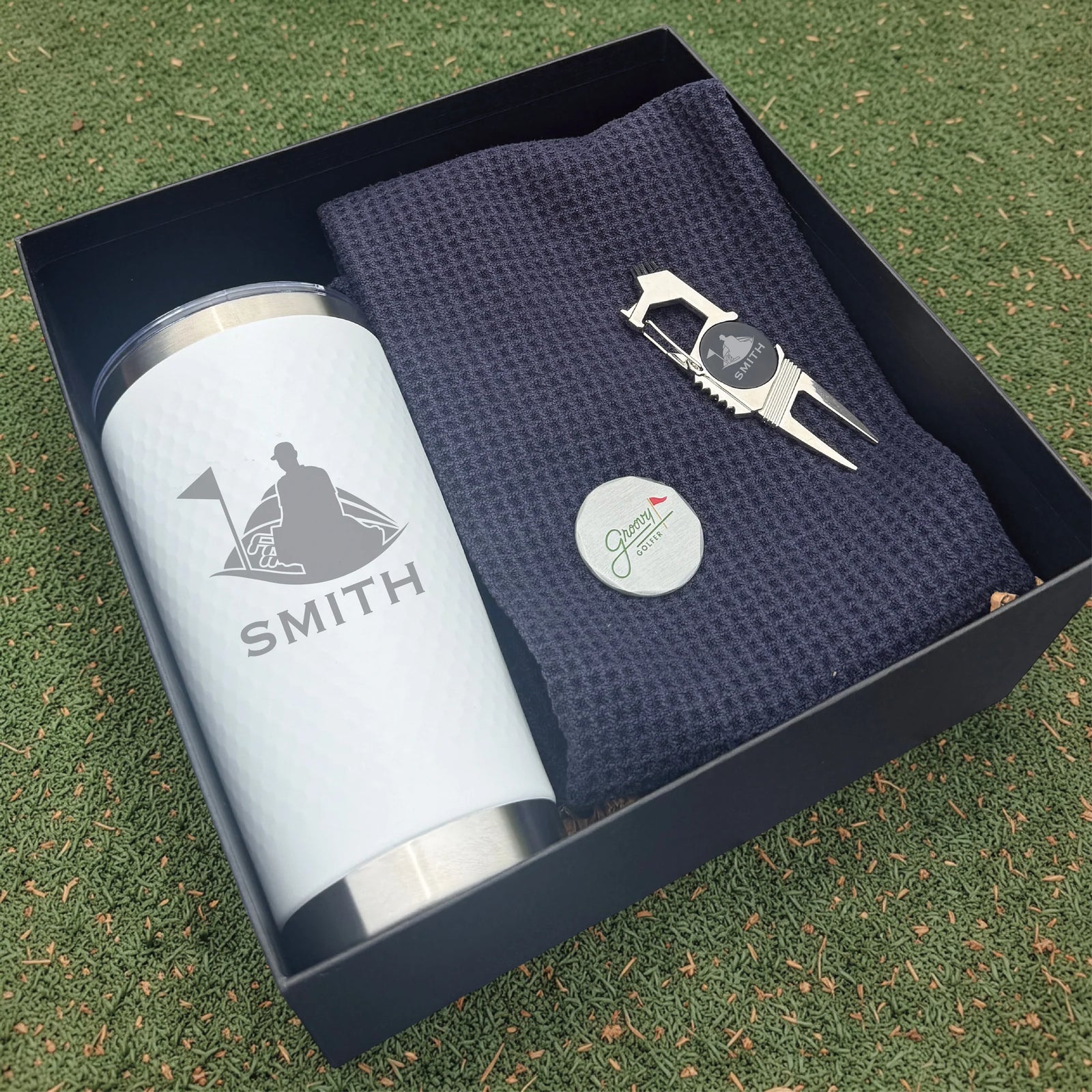 Golf-themed gift set with tumbler, towel, and divot tool in a box on grass.