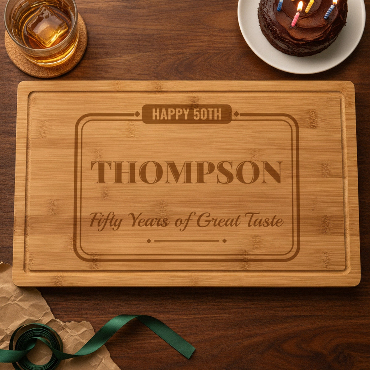 Personalized 50th birthday cutting board engraved Happy 50th, Thompson and Fifty Years of Great Taste beside a chocolate birthday cake, bourbon and unwrapped gift paper