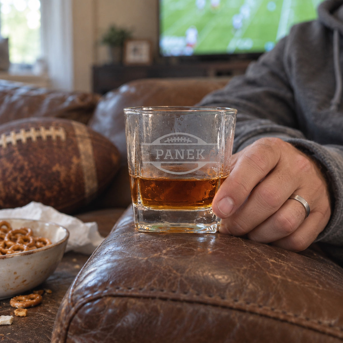 A man rests his personalized Bourbon Blitz football whiskey glass on the couch arm during a Sunday football game at home