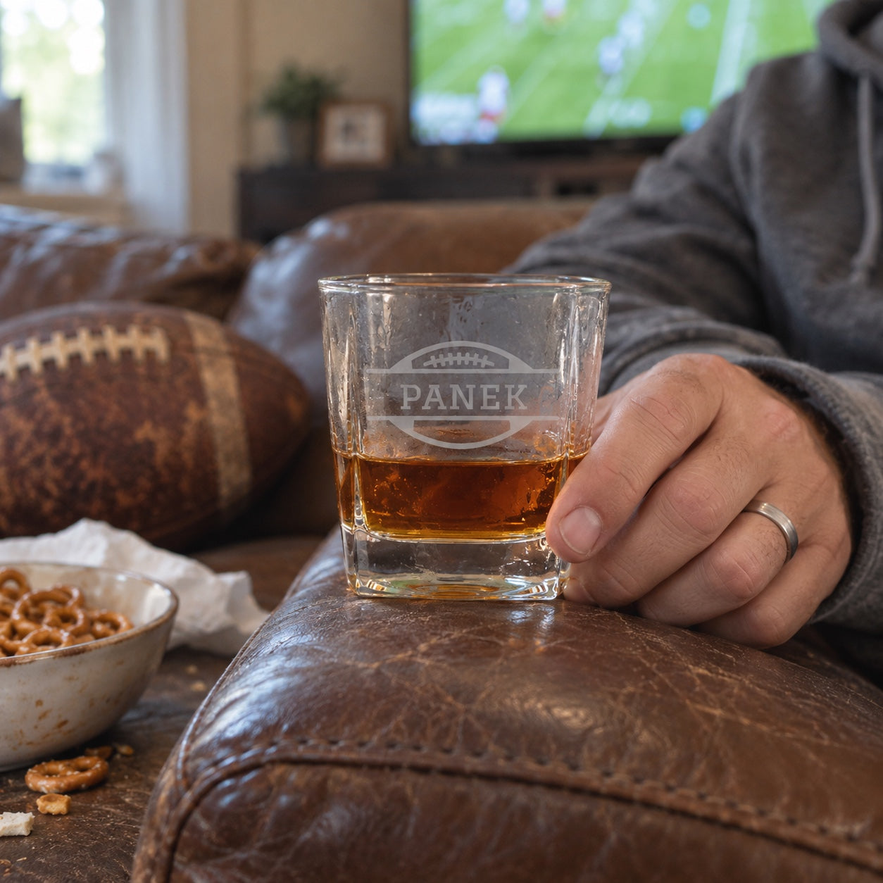 A man rests his personalized Bourbon Blitz football whiskey glass on the couch arm during a Sunday football game at home
