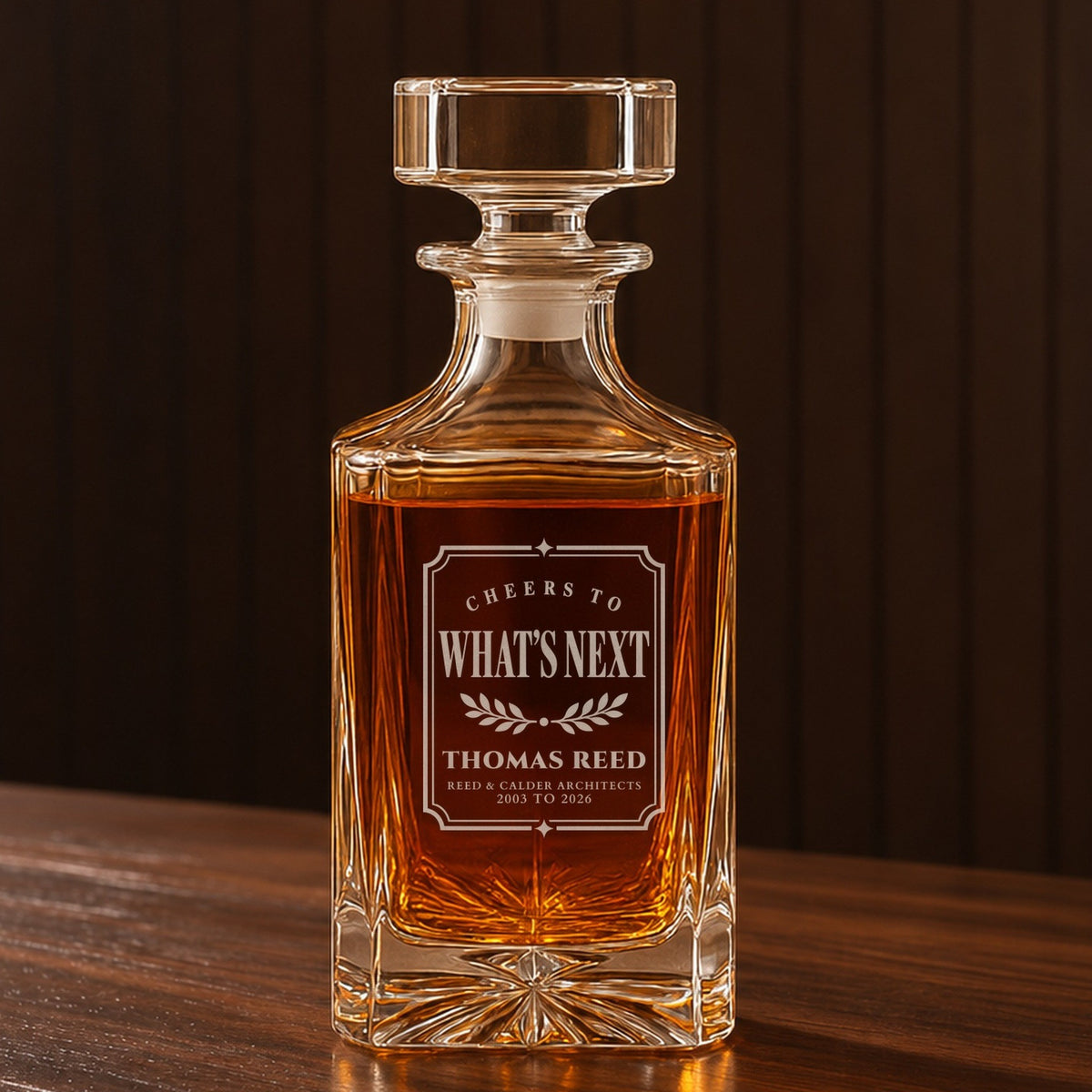 Personalized whiskey decanter with a framed Cheers to What's Next label engraved for Thomas Reed and his architecture firm, on a walnut bar