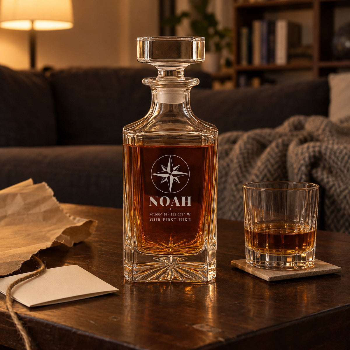 Personalized decanter for boyfriend on a coffee table beside torn kraft wrapping paper, engraved Noah with Seattle coordinates and Our First Hike