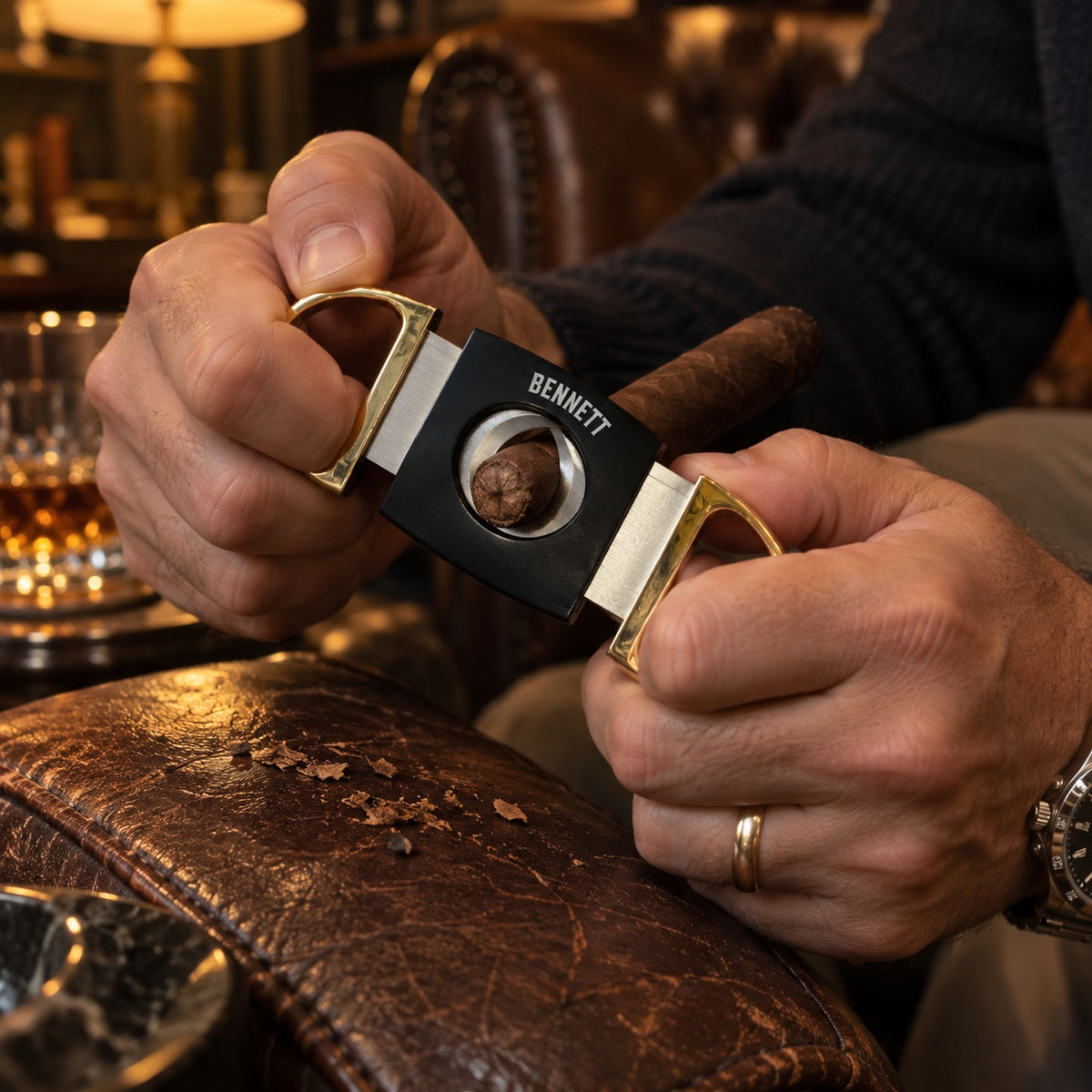 A man clips the cap off a cigar with his personalized black and gold cigar cutter engraved BENNETT, sitting in a leather chair in a cigar lounge.