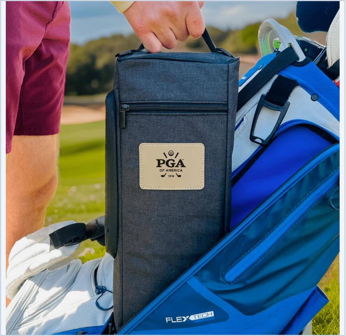 Dark gray golf cooler bag with PGA of America leather patch, held by golfer on course with golf bag and beverages visible