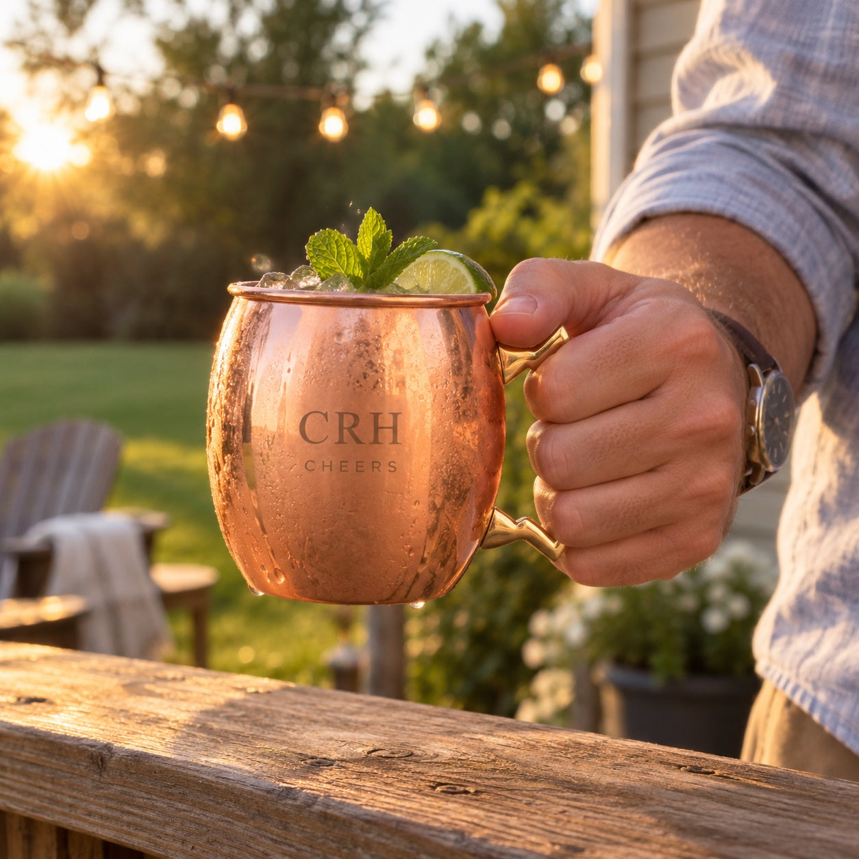 A man lifts his engraved copper Moscow mule mug with the initials CRH and CHEERS off a porch railing at golden hour, string lights and lawn behind.