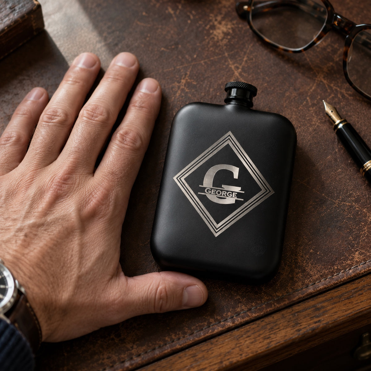 Custom engraved hip flask in matte black with a diamond monogram of a large G and the name George, on a leather desk beside a man's hand