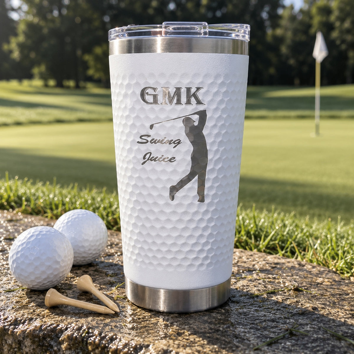 Custom golf ball tumbler with a dimpled white finish engraved GMK, Swing Juice and a swinging golfer, beside golf balls by the green