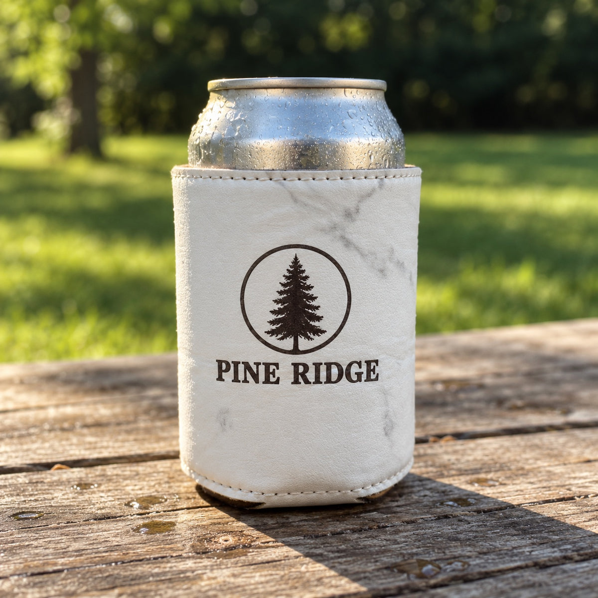 White marbled faux leather custom logo koozie engraved in dark brown with a company logo, holding a 12 oz can on a picnic table