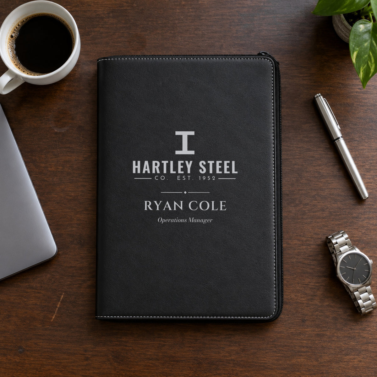 Custom logo padfolio in black, engraved in silver with a sample Hartley Steel logo, the name Ryan Cole and Operations Manager on a walnut desk