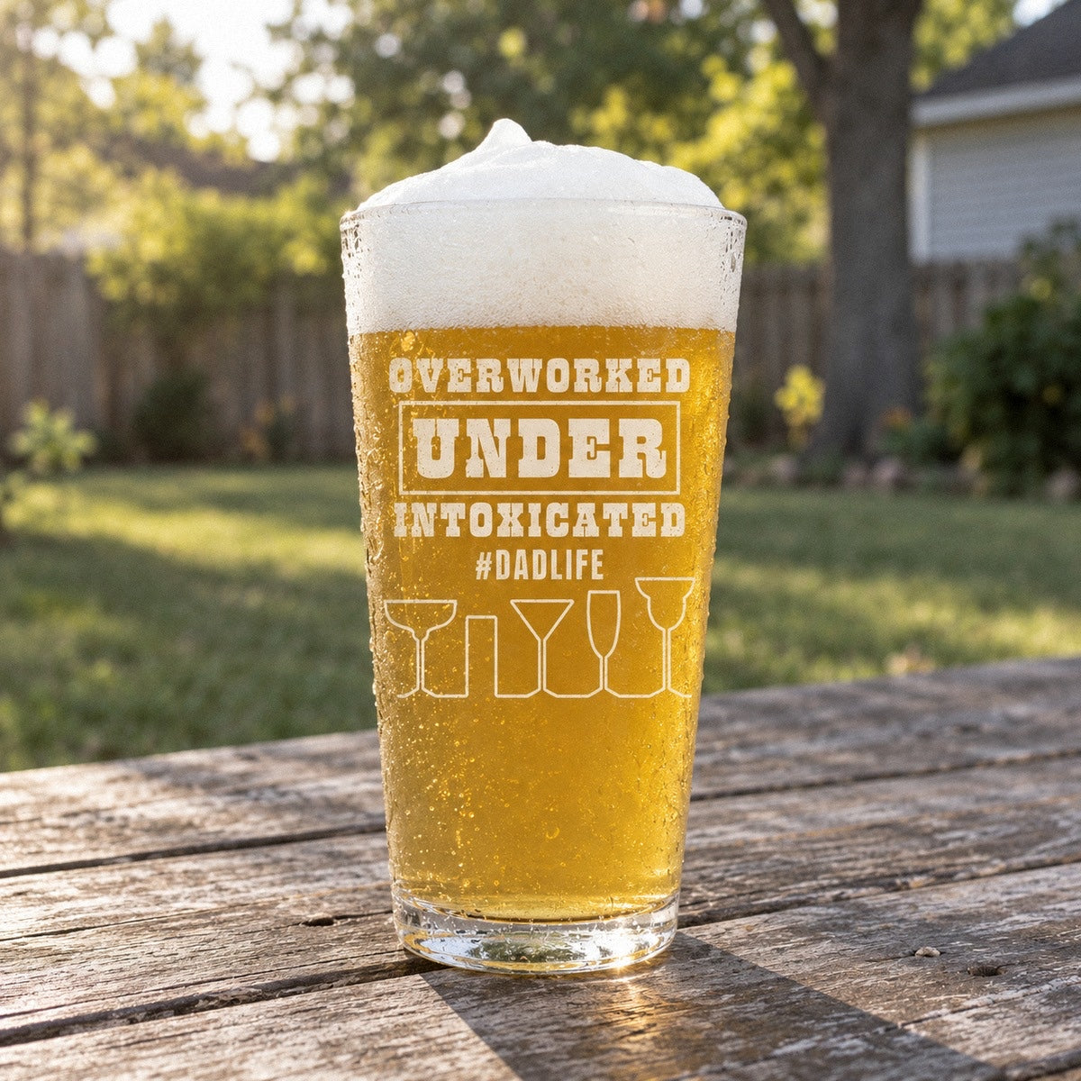 Dad beer glass full of lager on a porch table, printed with Overworked, Under, Intoxicated and #DADLIFE in bold white letters
