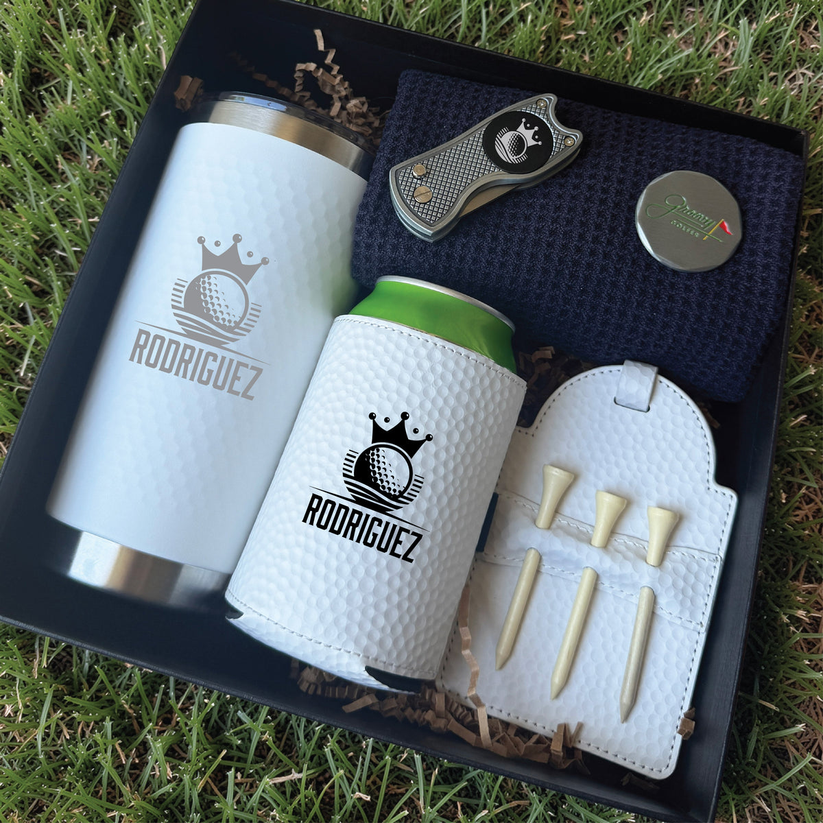 Golf gift set in black box containing white insulated tumbler and can cooler engraved with sample name "RODRIGUEZ" and crown logo, divot tool, tee holder with wooden tees, and metal ball marker on navy fabric.
