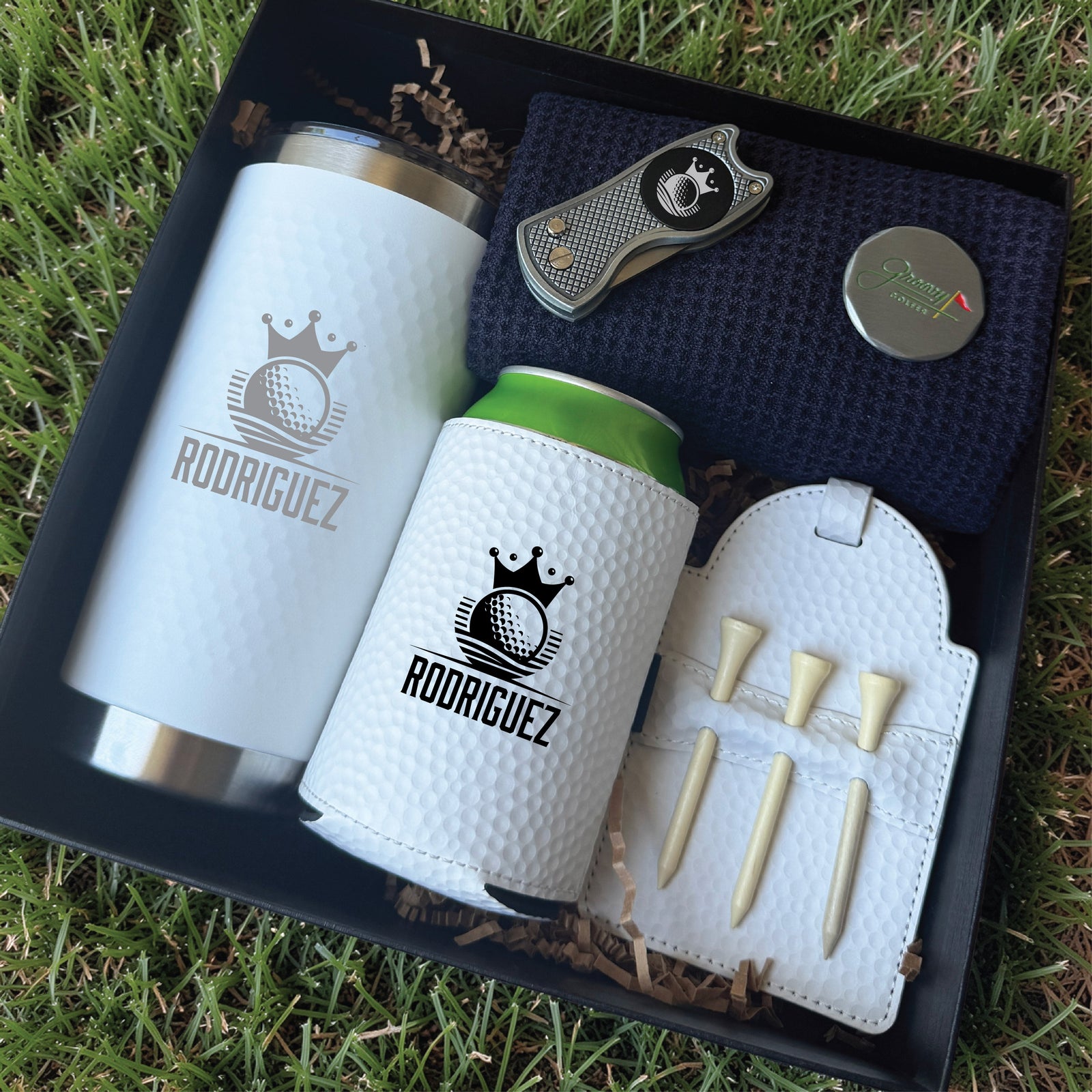 Golf gift set in black box containing white insulated tumbler and can cooler engraved with sample name "RODRIGUEZ" and crown logo, divot tool, tee holder with wooden tees, and metal ball marker on navy fabric.