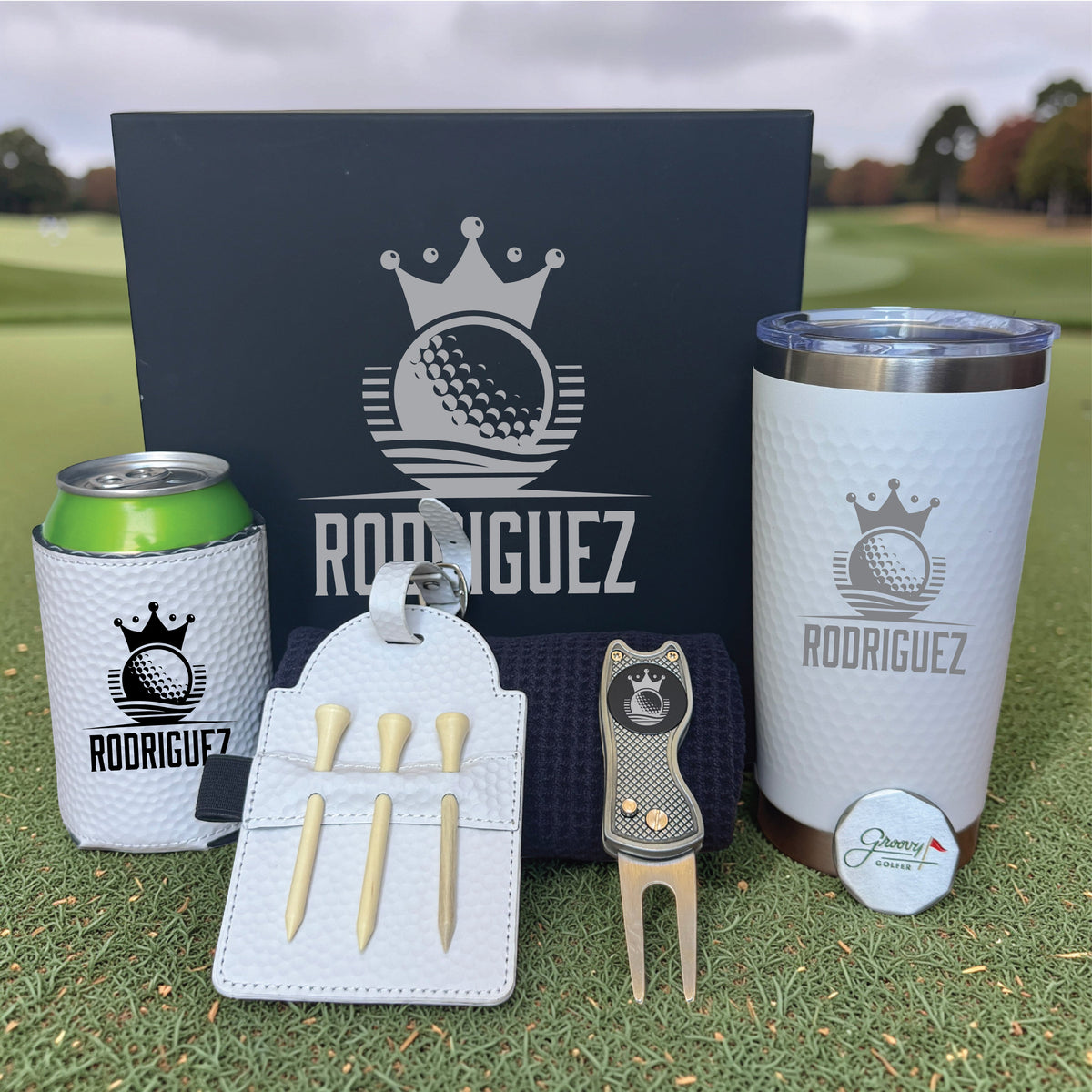 Golf set with personalized name "RODRIGUEZ" displayed on black gift box, white tumbler, can cooler, divot tool, tee holder with wooden tees, and ball marker on grass