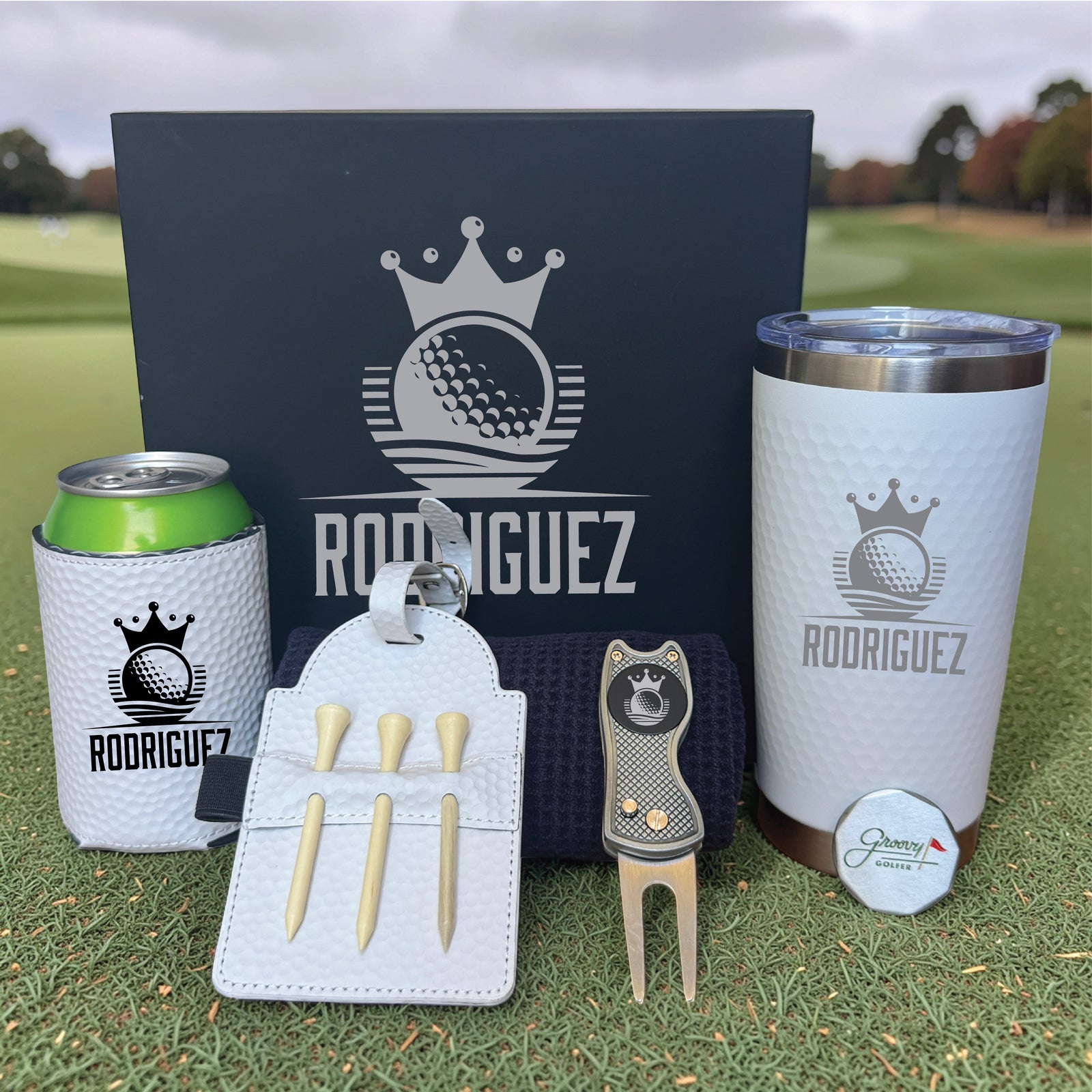 Golf set with personalized name "RODRIGUEZ" displayed on black gift box, white tumbler, can cooler, divot tool, tee holder with wooden tees, and ball marker on grass