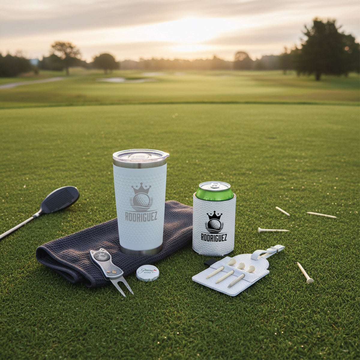 Personalized golf accessories displayed on putting green, including white tumbler and can cooler engraved with sample name "RODRIGUEZ" and crown logo, divot tool, ball marker, tee holder with wooden tees, and navy towel