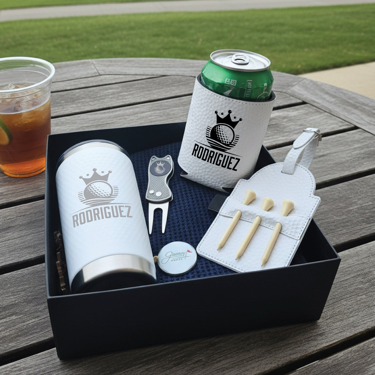 Golf gift set displayed in black box showing personalized tumbler, can cooler, divot tool, ball marker, and tee holder with sample "RODRIGUEZ" engraving and crown logo