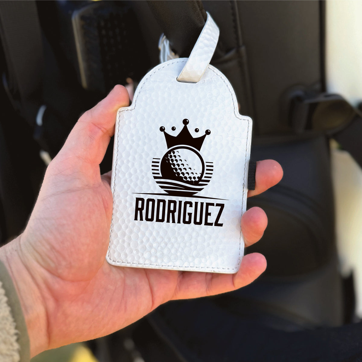 White leather golf towel tag with crown and golf ball motif, personalized with sample name "RODRIGUEZ" in black engraving, shown held in hand with white stitching detail and attachment loop