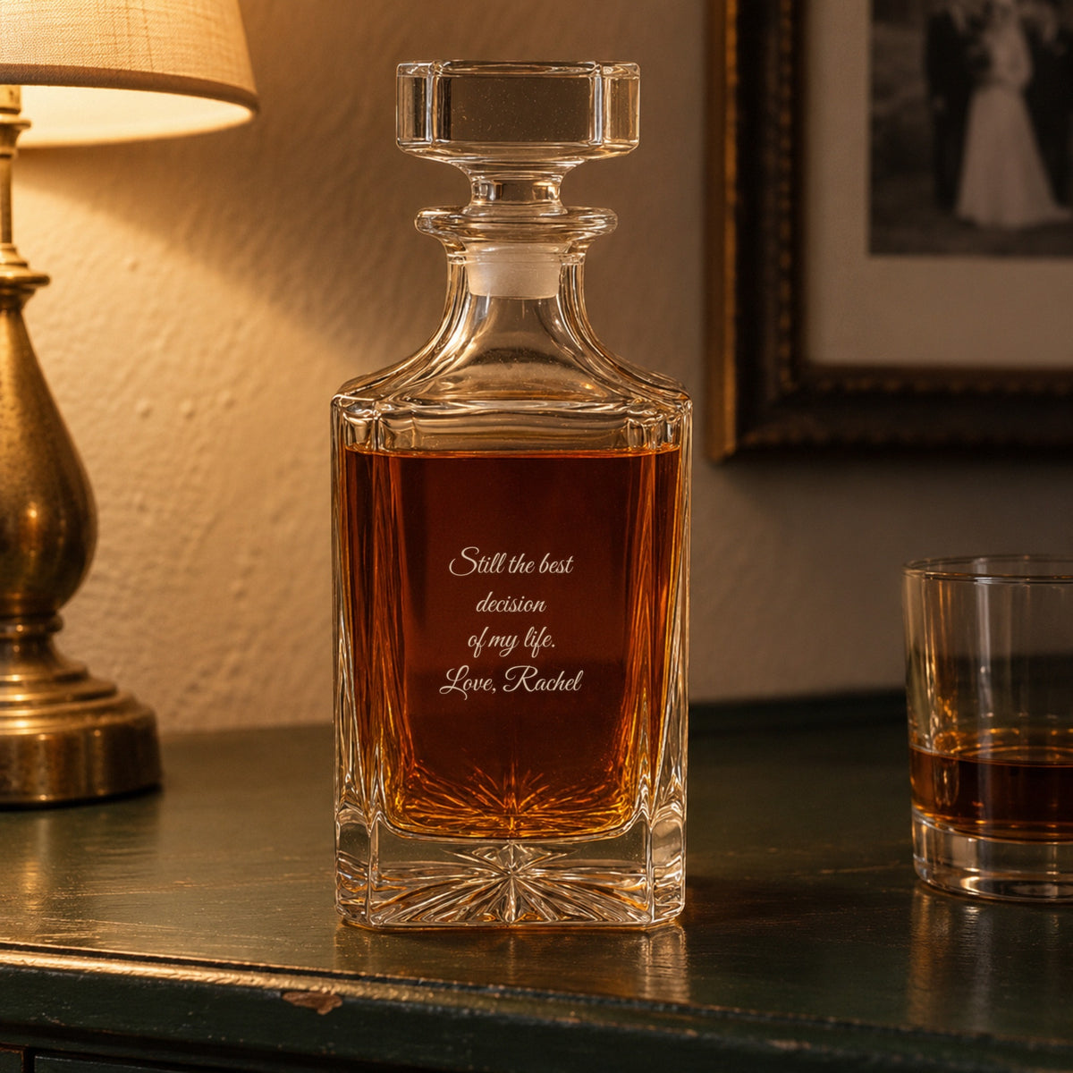 Back of a 15 year anniversary gift for him, the decanter engraved Still the best decision of my life, Love, Rachel, on a sideboard by a lamp