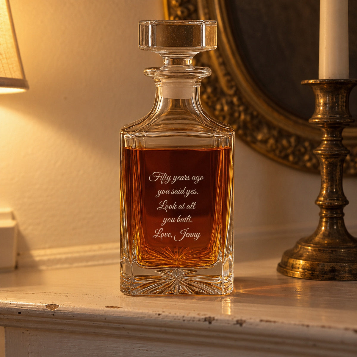 Back of a golden anniversary gift, the decanter engraved Fifty years ago you said yes, look at all you built, Love, Jenny, on a mantel by a candlestick