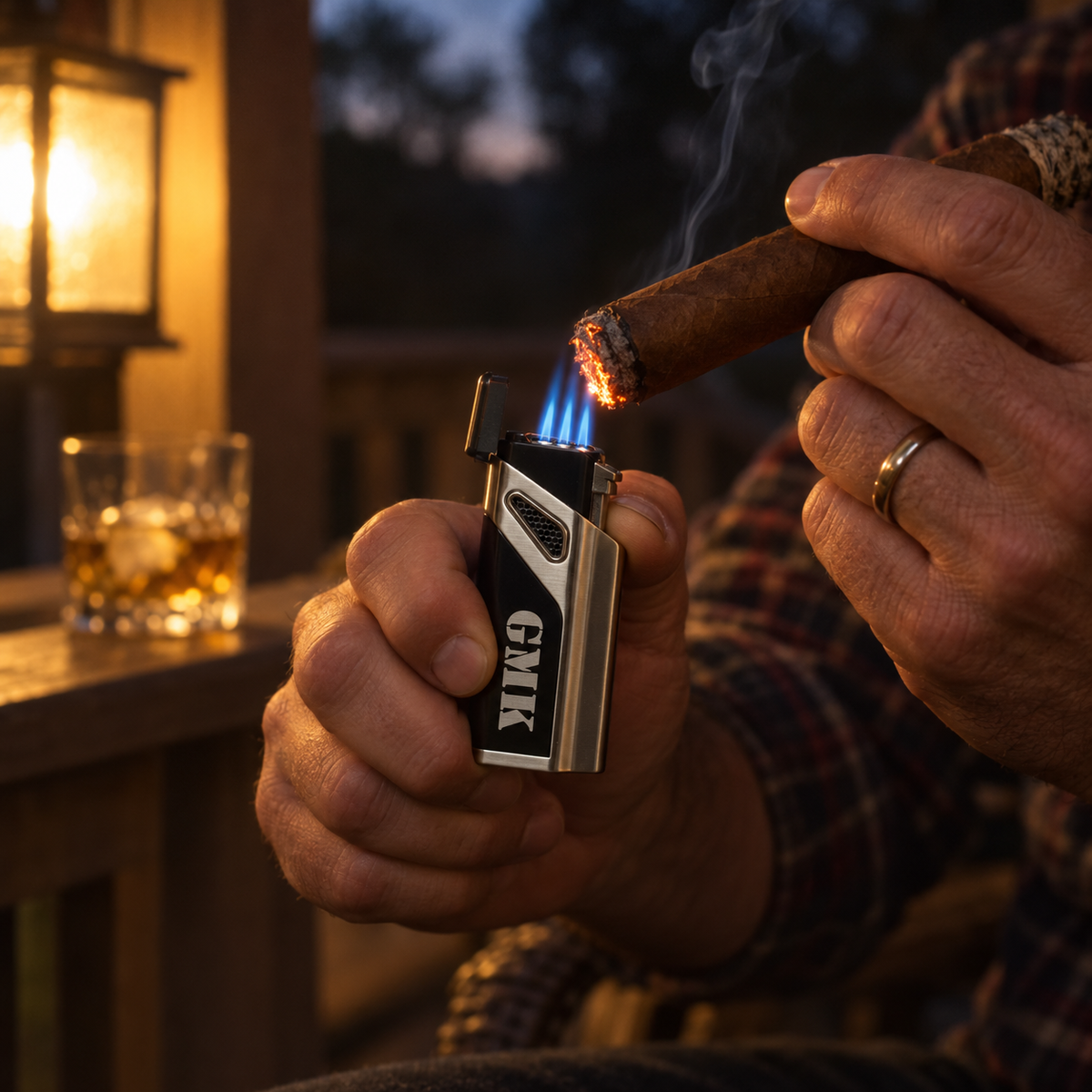 Engraved cigar lighter with GMK on the front lighting a cigar on a porch at dusk, its three blue torch flames lit, a glass of bourbon behind