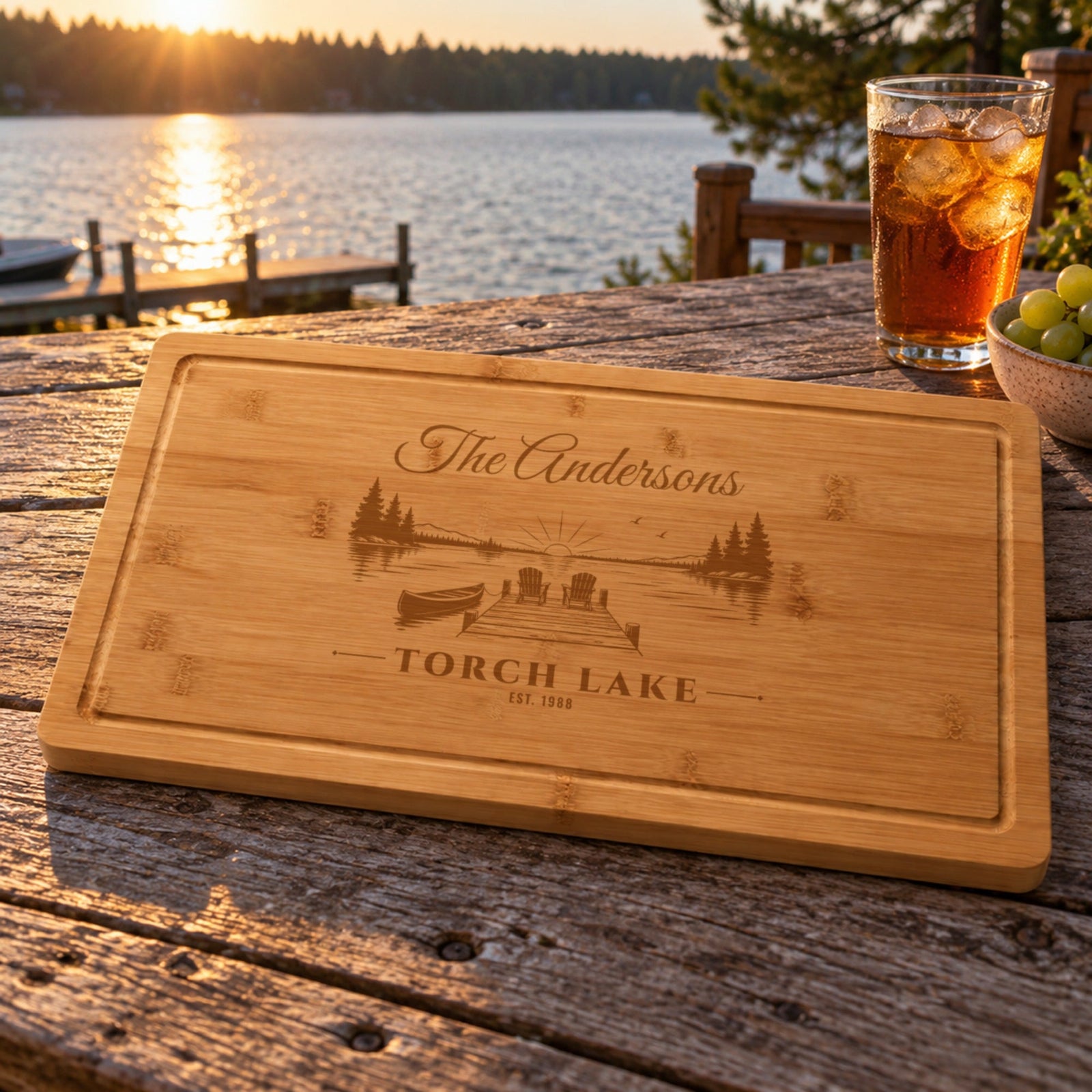 Engraved lake house gift: bamboo cutting board with The Andersons, Torch Lake, EST. 1988 and a dock scene, on a deck table by the lake at sunset