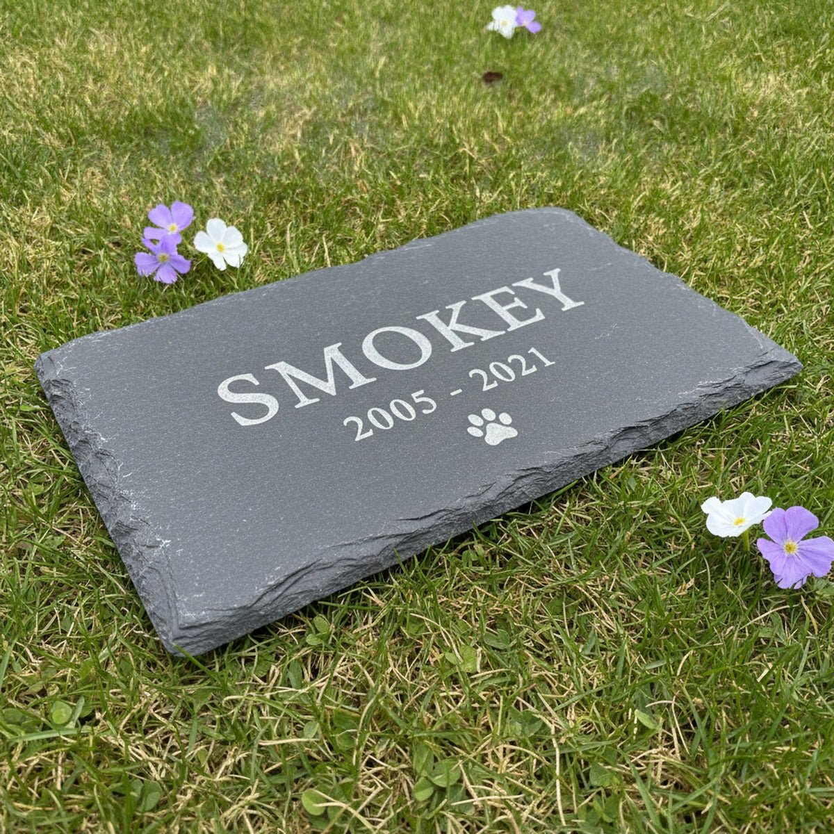 Custom slate pet memorial marker lying flat on garden grass among small purple and white flowers, engraved with the sample name Smokey, the years 2005 to 2021 and a paw print