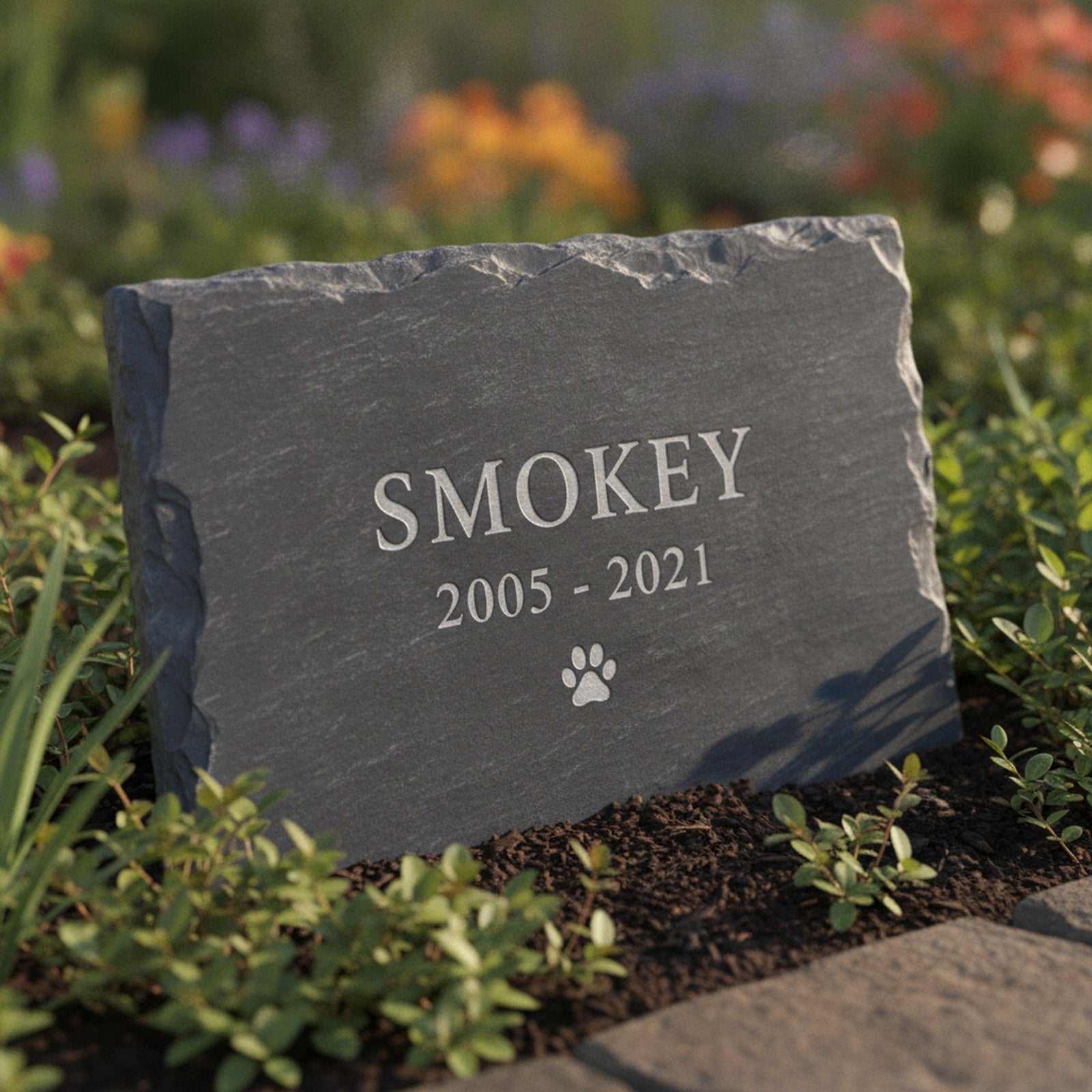 Personalized slate pet grave marker standing in a garden bed, laser engraved with the sample name Smokey, the years 2005 to 2021 and a small paw print