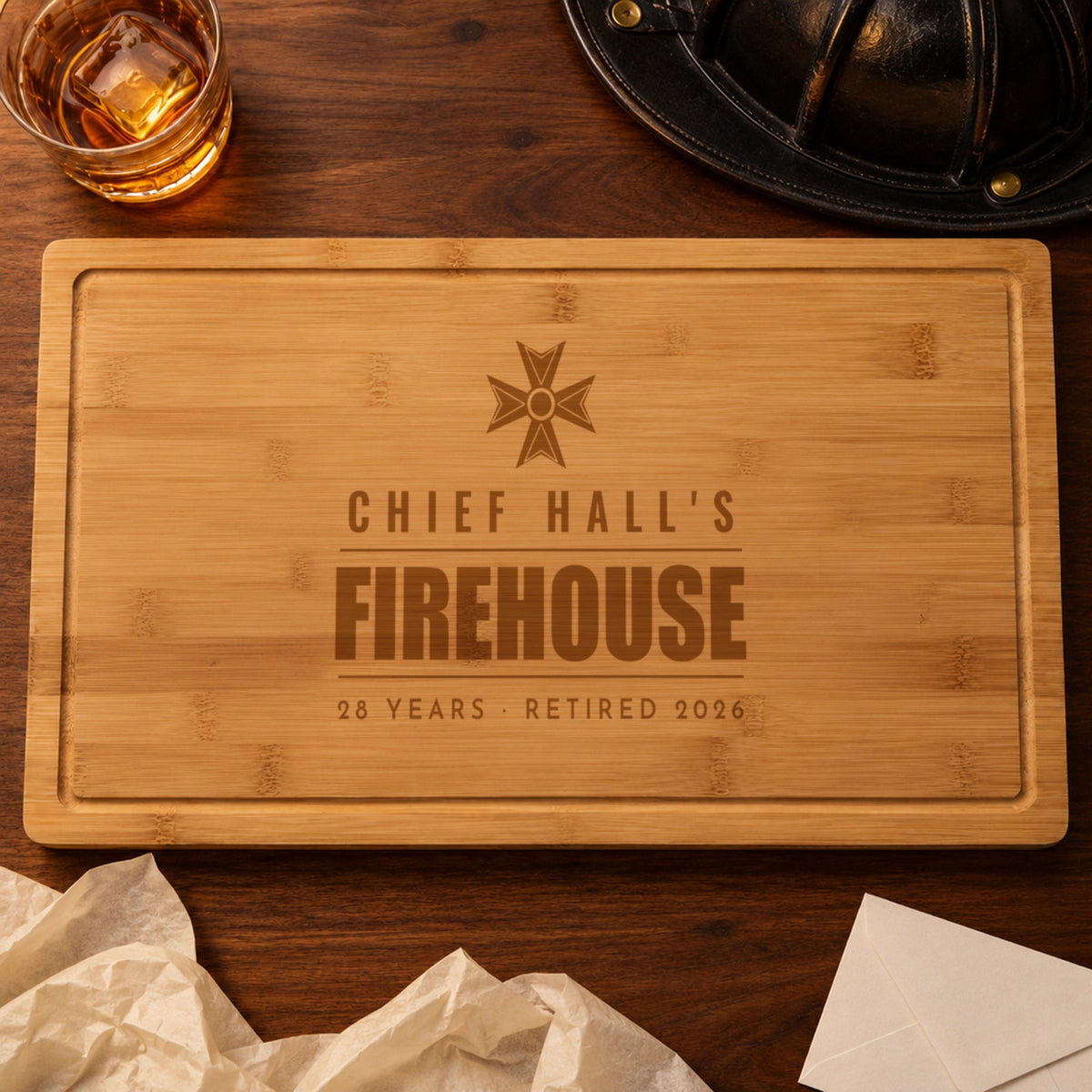 Firefighter retirement gift: bamboo cutting board engraved CHIEF HALL'S FIREHOUSE 28 YEARS RETIRED 2026 beside a black fire helmet and whiskey