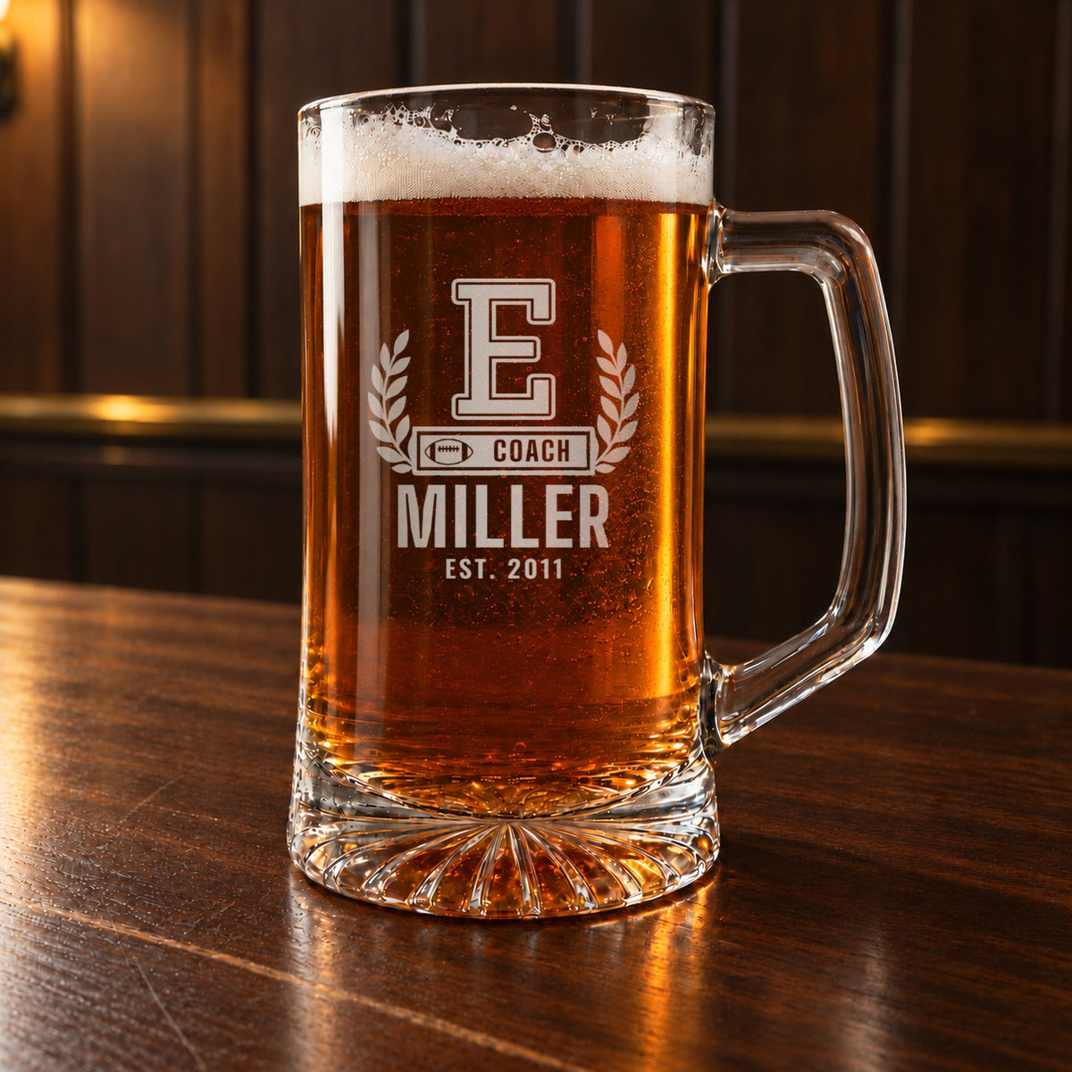 Personalized football coach letterman beer stein on a walnut bar, engraved with a varsity letter with laurel and a COACH bar: E, Coach, Miller, Est. 2011