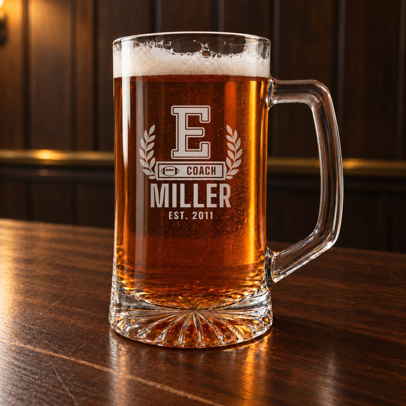 Personalized football coach letterman beer stein on a walnut bar, engraved with a varsity letter with laurel and a COACH bar: E, Coach, Miller, Est. 2011