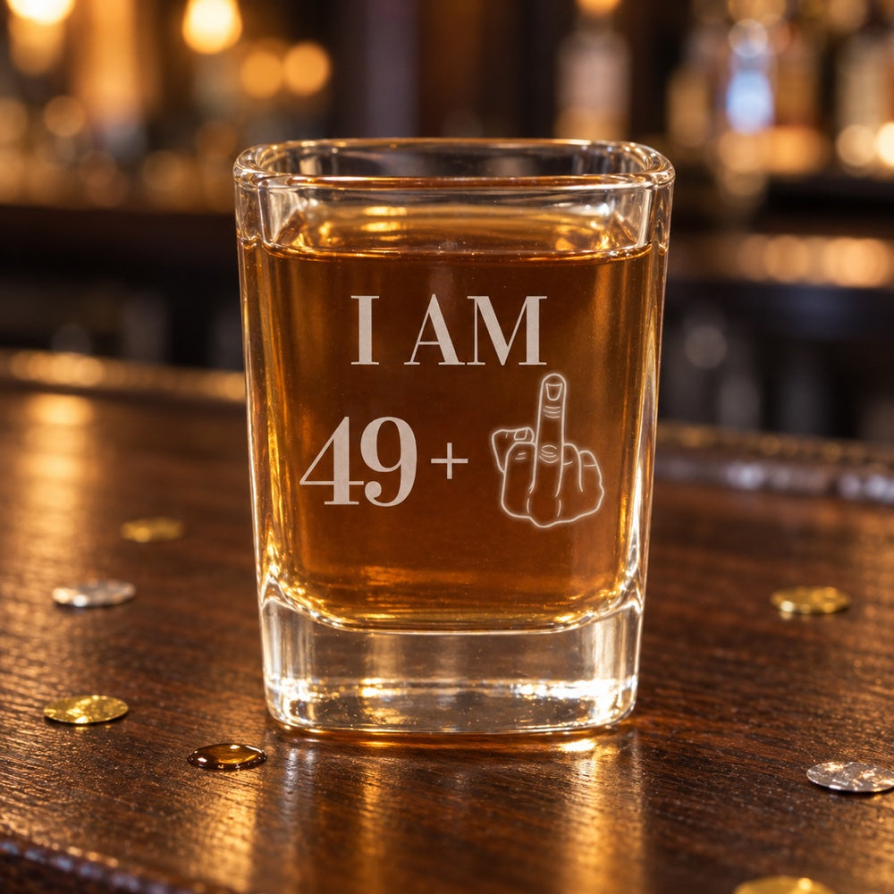 Funny 50th birthday shot glass engraved I AM 49 plus a middle finger, filled with whiskey on a dark walnut bar with gold and silver confetti