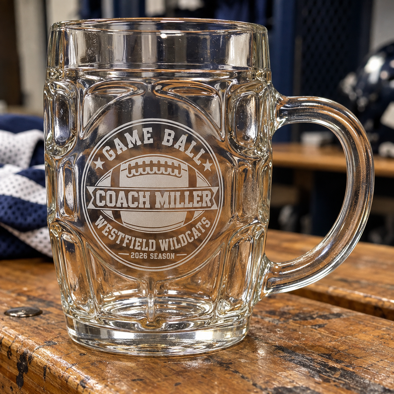 Game Ball personalized football coach beer stein on a locker room bench, frosted engraving reading Coach Miller, Westfield Wildcats, 2026 Season