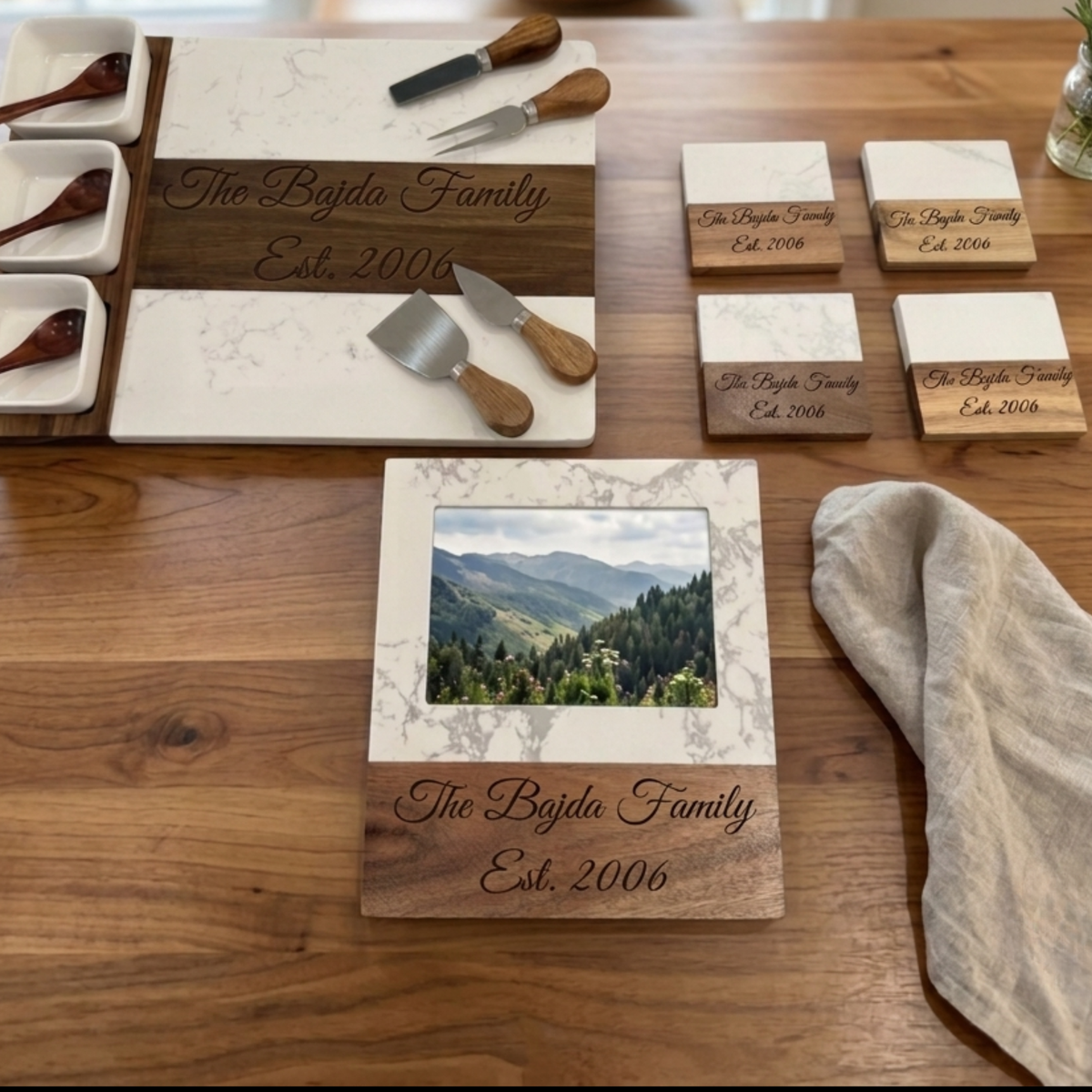 Personalized housewarming gift set for men laid out on a wood table with a marble and acacia charcuterie board, four matching coasters and a picture frame, each engraved with a family name and year.