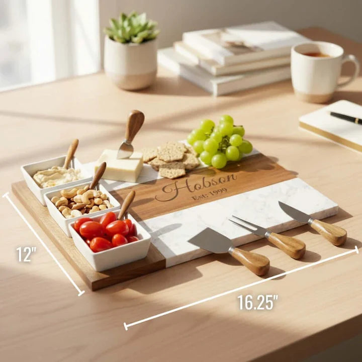 Marble and acacia charcuterie board with a sample name engraving, shown with white ceramic dipping bowls, spreaders, cheese, crackers and grapes ready for guests.