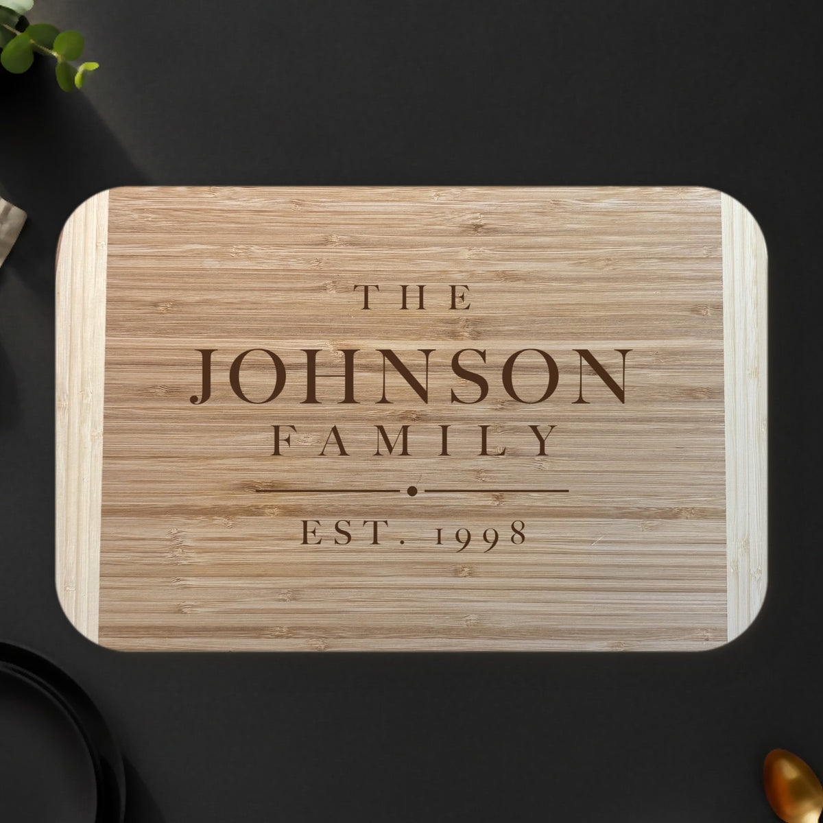 Personalized family name cutting board on a dark charcoal stone counter with eucalyptus and lemons, engraved THE JOHNSON FAMILY EST. 1998 on two tone bamboo.