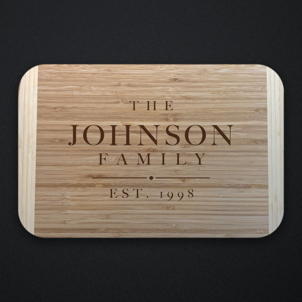 Two tone bamboo cutting board engraved THE JOHNSON FAMILY with EST. 1998 beneath a rule and centre dot, shown on a dark slate background.