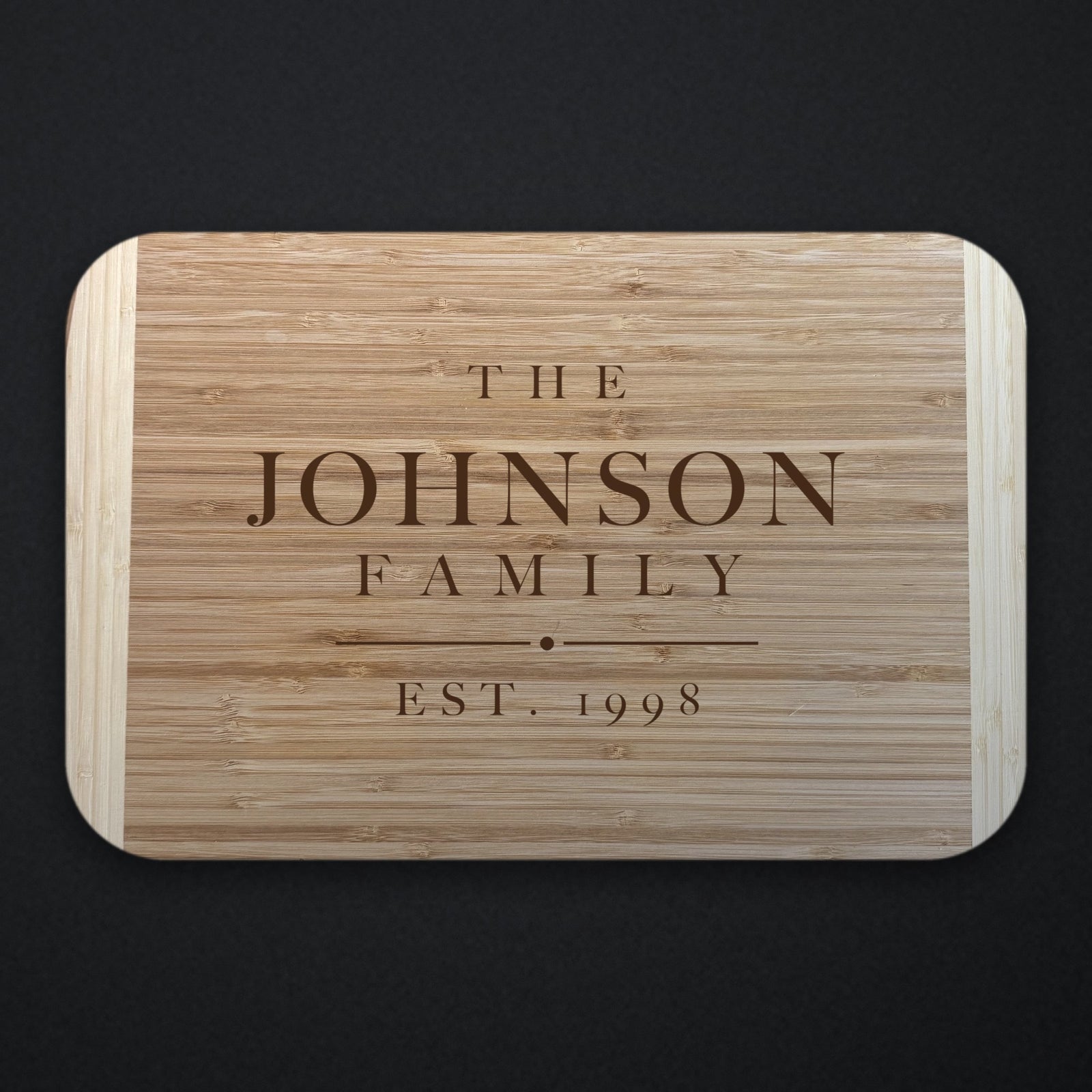 Two tone bamboo cutting board engraved THE JOHNSON FAMILY with EST. 1998 beneath a rule and centre dot, shown on a dark slate background.