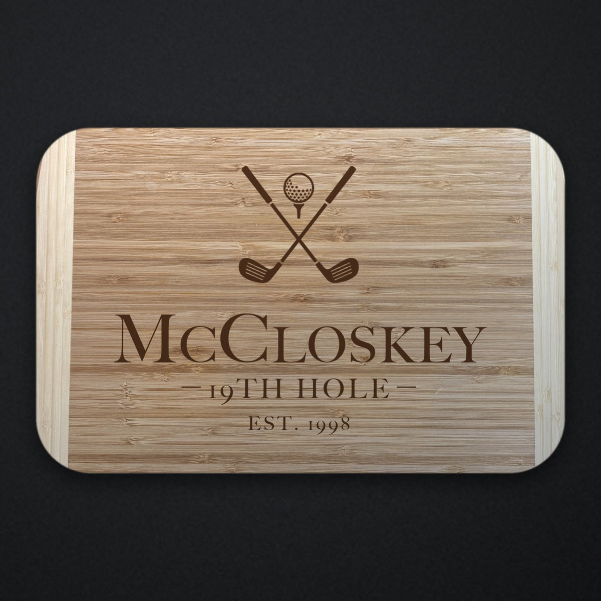 Two tone bamboo cutting board engraved with crossed golf clubs and a ball above the name McCloskey, the line 19TH HOLE and EST. 1998, shown on a dark slate background.