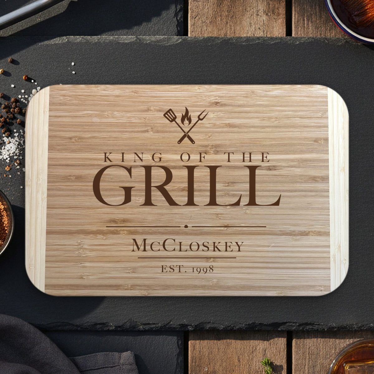 Personalized King of the Grill cutting board on dark slate with grilling tongs, dry rub and bourbon, engraved McCloskey EST. 1998 under crossed grill tools on two tone bamboo.