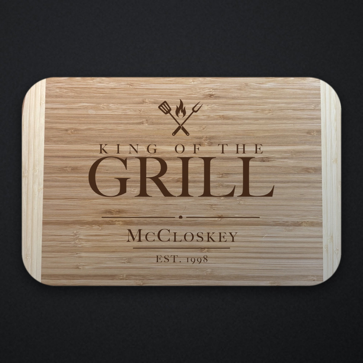 Two tone bamboo cutting board engraved KING OF THE GRILL under crossed grilling tools and a flame, with the name McCloskey and EST. 1998 below, on a dark slate background.
