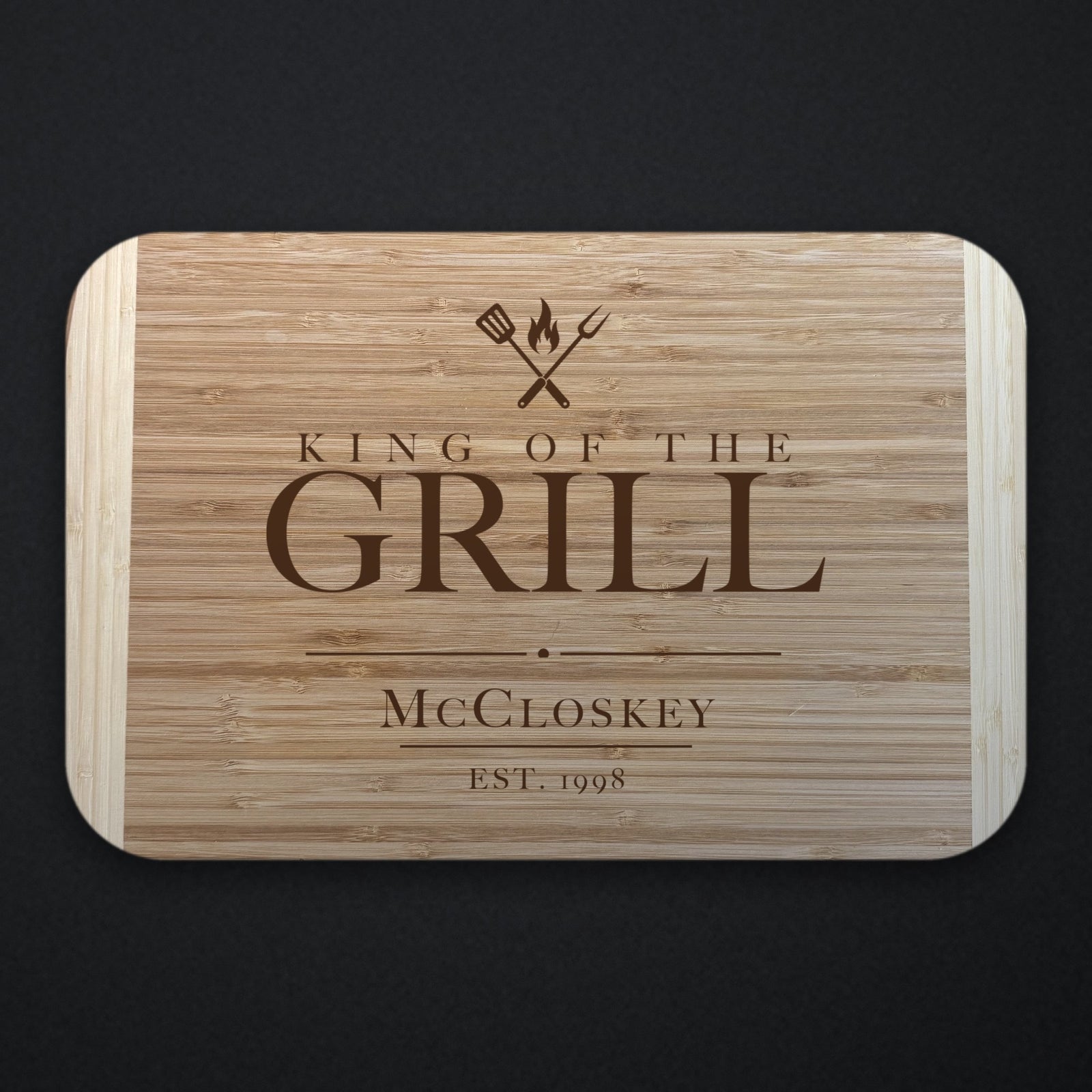 Two tone bamboo cutting board engraved KING OF THE GRILL under crossed grilling tools and a flame, with the name McCloskey and EST. 1998 below, on a dark slate background.